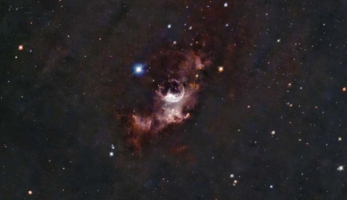 The Bubble Nebula — a bright curved shell of gas inside a cloud of rust-red nebula, a blue star to its upper left, in a field of small stars